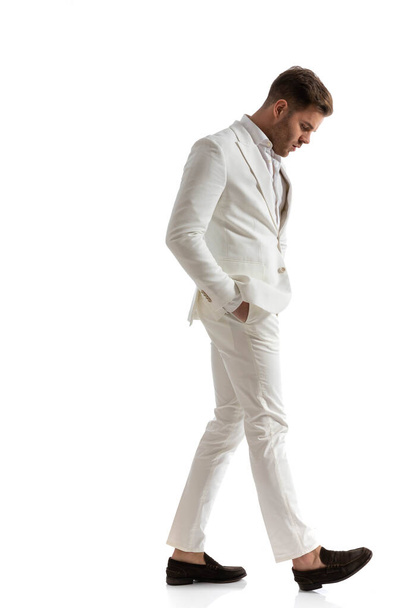 白いスーツを着た魅力的なエレガントな男がポケットに手を取り歩きスタジオの白い背景を見下ろし - 写真・画像