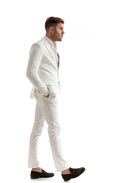 вид сбоку элегантного бизнесмена в белом костюме, держащего руки в карманах и идущего изолированно на белом фоне в студии - Фото, изображение
