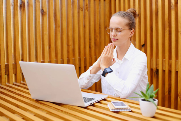 Horizontal shot of relaxed Caucasian woman with hair bun wearing white shirt and optical eyeglasses sitting and working on laptop, doing yoga practice for calming down and concentrate. - Foto, Bild