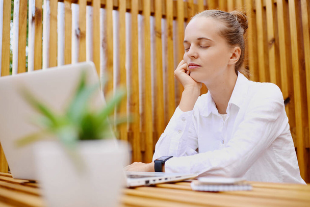 Horizontal shot of bored tired woman with hair bun wearing white shirt sitting and working on laptop, posing with closed eyes, keeps hand under chin, relaxing during break. - Фото, изображение