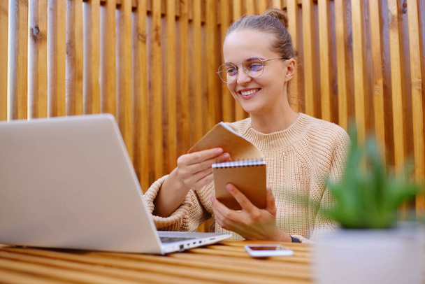 Horizontal shot of attractive winsome young adult woman with bun hairstyle wearing beige sweater working on laptop, holding paper organizer, smiling happily. - Foto, Imagem