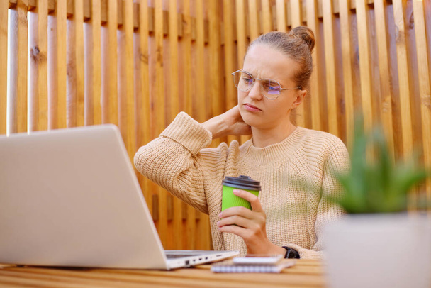 Image of tired exhausted woman with bun hairstyle wearing beige sweater working on laptop and drinking coffee, feels terrible pain in neck, works during long hours. - Фото, изображение