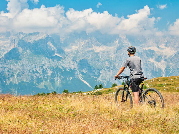 Teenager sit on modern mtb and watching from hill peak to opposite stonny and snowy peaks. Dolomite Alps, Trento region, Italy  - Photo, Image