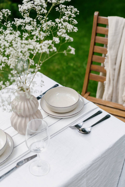 slavnostní oběd venku v létě, stůl s bílým plátnem ubrus, prázdný talíř a miska, víno sklo, vidlička, lžička a stříbrný nůž, váza s květinami v zelené zahradě - Fotografie, Obrázek