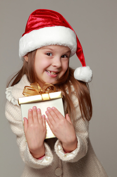 Pretty smiling little girl in Santa's red hat holding Christmas box - Фото, изображение