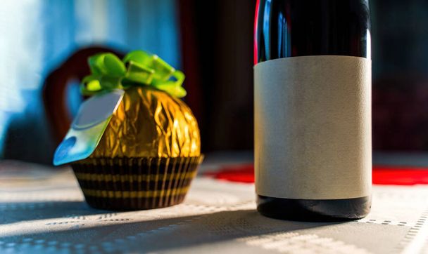 Κοντινό πλάνο ενός κόκκινου μπουκαλιού κρασιού με μαύρη λευκή ετικέτα για mock up με μια μεγάλη στρογγυλή τυλιγμένη σοκολάτα σε χρυσό σε ένα τραπέζι, δώρο και εορταστική ιδέα κατά τη διάρκεια του φεστιβάλ. - Φωτογραφία, εικόνα