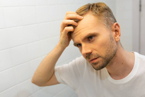 Biały mężczyzna w średnim wieku z krótką brodą patrzy na włosy w lustrze w łazience i martwi się łysieniem. Koncepcja problemu wypadania włosów u mężczyzn, wczesnej łysienia i łysienia. - Zdjęcie, obraz