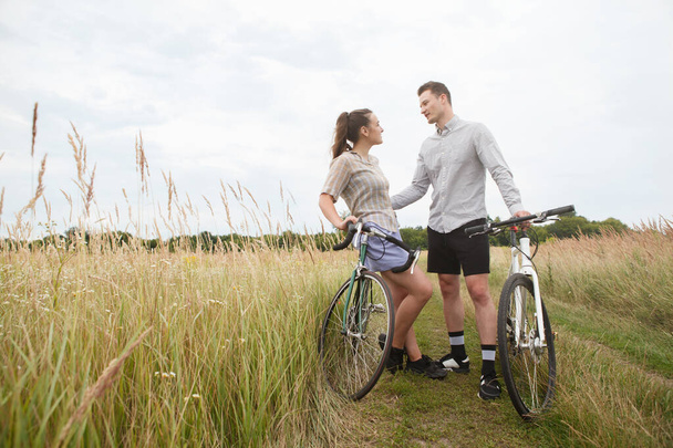 フィールドの近くの幸せなカップルサイクリング。自転車のある男と女は夏に畑の近くに行く. - 写真・画像