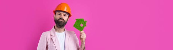 Retrato de un joven ingeniero de construcción que usa un casco naranja, con una chaqueta rosa sobre un fondo rojo de estudio. Un hombre tiene una eco casa verde. - Foto, Imagen