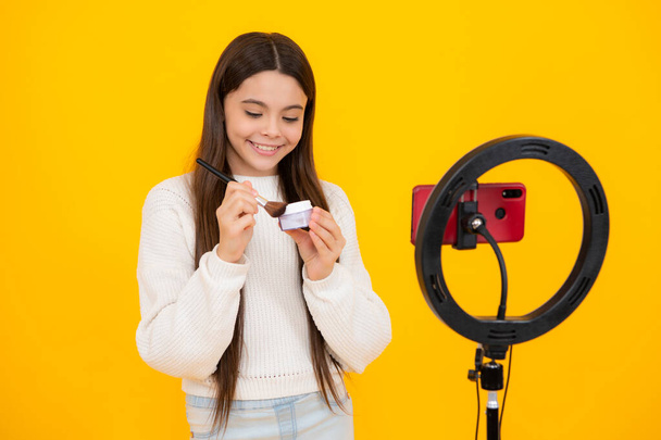 Beauty blog, presenting makeup cosmetics powder and brush. Teenager child blogger with phone recording video on isolated yellow studio background. Influencer teen girl speaking in front of smartphone - Photo, Image