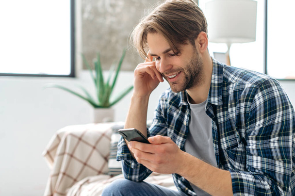 Fröhlicher kaukasischer attraktiver Kerl, der sein Smartphone benutzt, während er zu Hause auf dem Sofa sitzt, im Internet und in sozialen Netzwerken surft, mit Freunden oder Familie SMS schreibt, gute Nachrichten erfährt, glücklich lächelt - Foto, Bild
