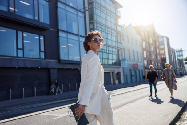Красива жінка в модному вбранні перетинає дорогу на тлі міських будівель і дивиться в сторону
 - Фото, зображення