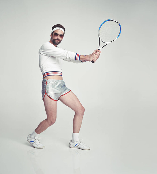 A young man in the studio wearing ill-fitting retro tennis wear and shades while holding a racquetA young man in the studio wearing ill-fitting retro tennis wear and shades while holding a racquet. - Fotoğraf, Görsel