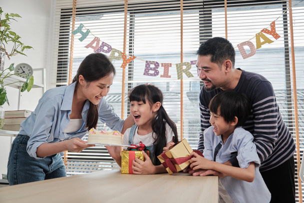 Happy Asian Thai rodziny, młoda słodka dziewczyna zaskoczona tort urodzinowy, prezent, świeca dmuchać, i świętować imprezę z rodzicami i rodzeństwem razem przy stole, dobre samopoczucie domu specjalne wydarzenie. - Zdjęcie, obraz