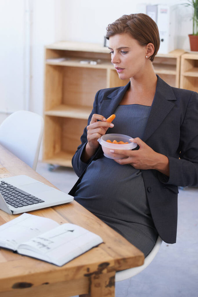 Giving in to pregnancy cravings. A pretty and pregnant businesswoman satisfying a craving while looking at her laptop screen - Φωτογραφία, εικόνα