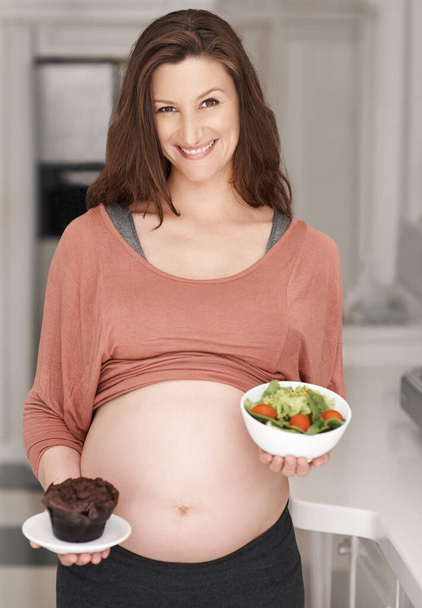 Abbiamo un vincitore. Ritratto ritagliato di una giovane donna incinta che decide tra un'insalata o un cupcake in cucina - Foto, immagini