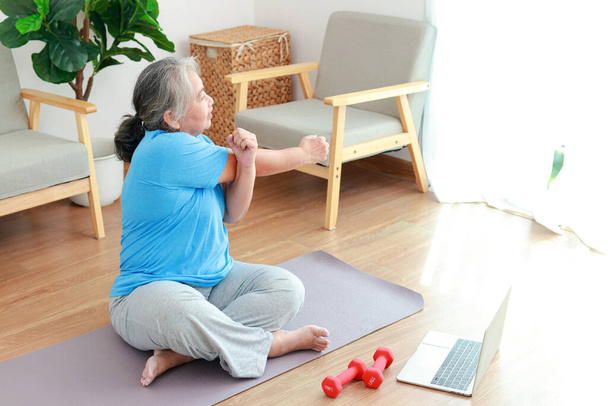 Asian elderly woman sitting at home exercising, doing exercises according to online fitness trainers. through a video call on a laptop. Social distancing, maintaining the health of the elderly - Zdjęcie, obraz