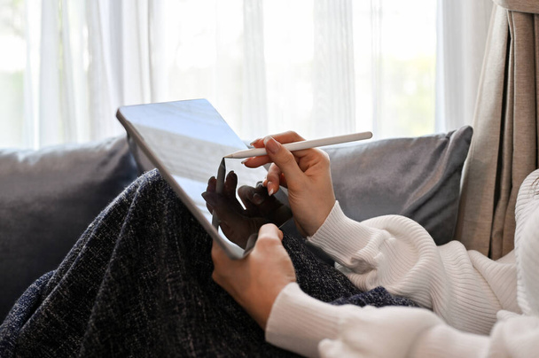 Közelkép és kivágott kép, Egy csinos nő ellazul a nappaliban, ül a kényelmes kanapén, és használ egy digitális tabletta touchpad. - Fotó, kép