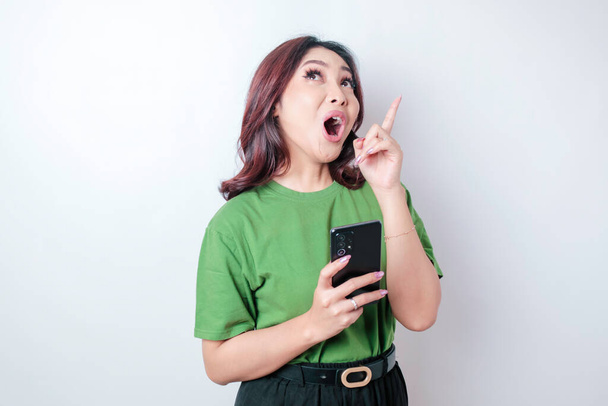 Shocked Asian woman wearing a green t-shirt pointing at the copy space upside her holding her phone, isolated by a white background - Foto, immagini