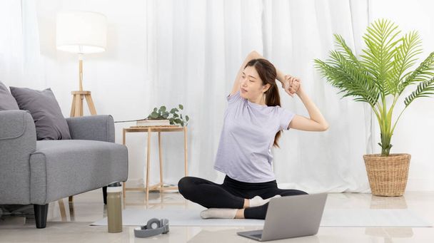 Beautiful Asian woman in sportswear is warming up or doing light exercises in the house with dumbbells and a protein shake or bottle of water and using a laptop at home, Healthy exercise. - Фото, изображение