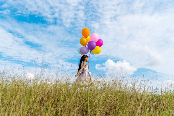 Wesoły cute girl gospodarstwa balony działa na zielonej łące białej chmury i błękitne niebo ze szczęściem. Dłonie trzymając żywe balony powietrza grać na urodziny happy times lato na słońcu zewnątrz - Zdjęcie, obraz