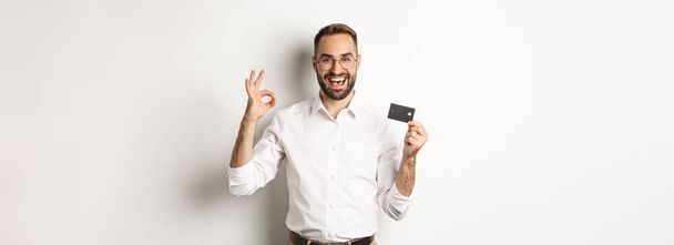 Schöner Mann mit Kreditkarte und Okay-Zeichen, Bankempfehlung, vor weißem Hintergrund stehend. Kopierraum - Foto, Bild