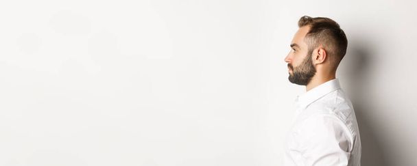 Close-up profile shot of handsome bearded man looking left, standing against white background. - Fotografie, Obrázek