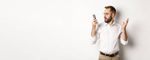 Mies katselee hämmentyneenä matkapuhelinta keskustelun jälkeen, seisoo ymmällään valkoisella taustalla. Kopioi tila - Valokuva, kuva