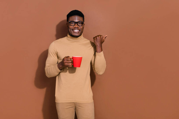 Πορτρέτο του όμορφου trendy χαρούμενος τύπος πίνοντας τσάι δείχνει διαφημιστικό χώρο αντίγραφο απομονωμένο πάνω από καφέ φόντο χρώμα. - Φωτογραφία, εικόνα