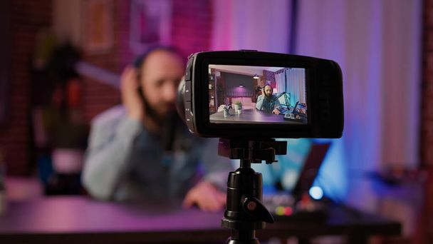 Selective focus on digital video camera screen recording male vlogger with headphones talking at microphone in home podcasting studio. Closeup of dslr filming influencer streaming live internet show. - Photo, Image