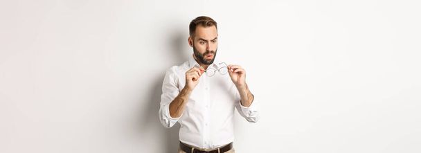 Hombre mirando confundido a sus gafas, de pie en ropa de oficina sobre fondo blanco. - Foto, imagen