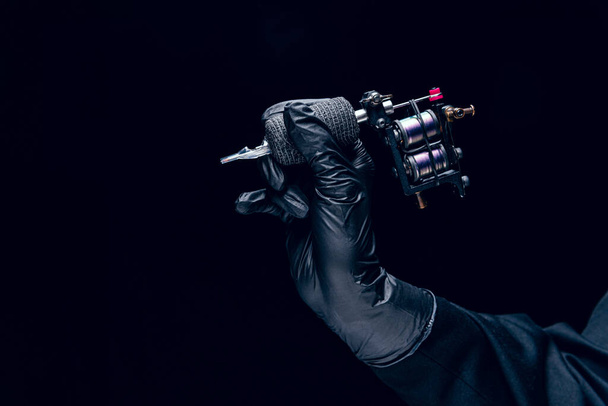 Machine for tattoo. Human hand in black protective glove holding professional equipment for making tattoo on body isolated on dark background. Concept of art, job, hobby, poster for ad - Фото, изображение