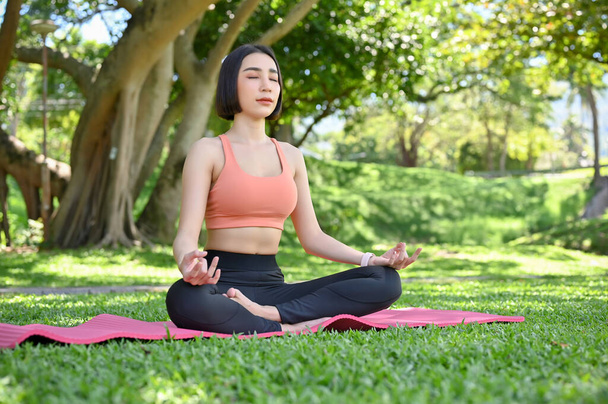 Güzel ve sakin genç Asyalı kadın spor kıyafetleriyle yoga yapıyor, derin derin nefes alıyor ve güzel yeşil bahçede meditasyon yapıyor. Lotus yoga pozu. Refah ve Meditasyon kavramı - Fotoğraf, Görsel
