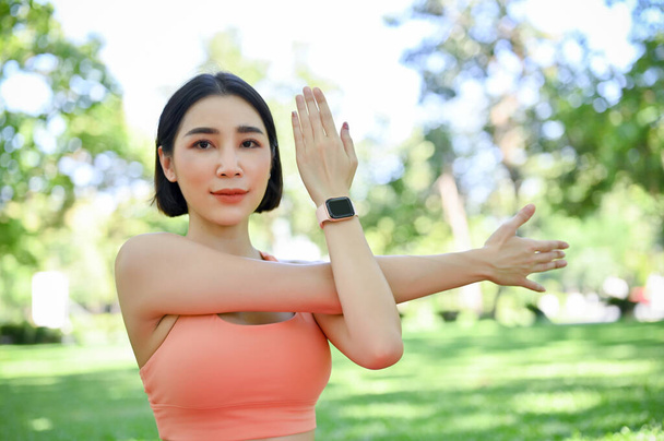 Ελκυστική νεαρή γυναίκα στα αθλητικά ασκεί γιόγκα στο πράσινο πάρκο, αναπνέει βαθιά και τεντώνει τα χέρια της. - Φωτογραφία, εικόνα