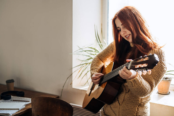 Уютные зимние хобби в помещении, осенний холодный сезон. Рыжая женщина играет на акустической гитаре и слушает музыку, сидит у окна дома. - Фото, изображение