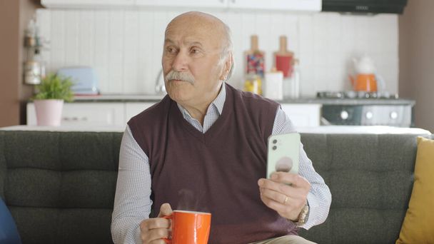 Γέρος βλέποντας αστείο βίντεο στο smartphone του ενώ έπινε καφέ στο σπίτι. Ο άνθρωπος γελάει με τα sms ή το βίντεο που βλέπει στο smartphone του και κοιτάζει τον άδειο διαφημιστικό χώρο. - Φωτογραφία, εικόνα