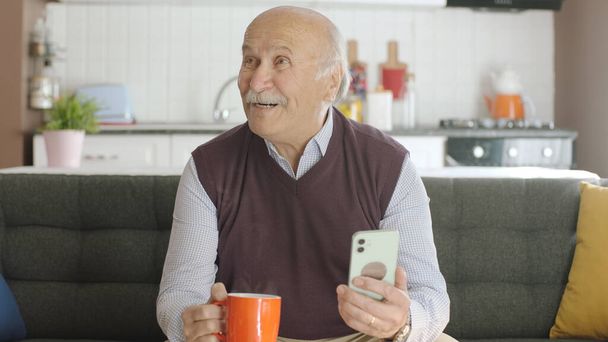 Alter Mann schaut lustige Videos auf seinem Smartphone, während er zu Hause Kaffee trinkt. Der Mann lacht über die SMS oder das Video, das er auf seinem Smartphone sieht, und blickt auf die leere Werbefläche. - Foto, Bild