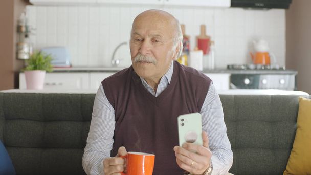 Viejo viendo videos divertidos en su teléfono inteligente mientras bebe café en casa. El hombre se ríe de los sms o video que ve en su teléfono inteligente y mira el espacio publicitario vacío. - Foto, imagen