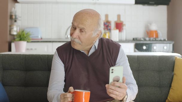 Viejo viendo videos divertidos en su teléfono inteligente mientras bebe café en casa. El hombre se ríe de los sms o video que ve en su teléfono inteligente y mira el espacio publicitario vacío. - Foto, imagen