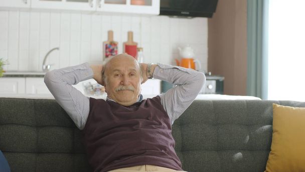 高齢者の男はアームチェアに寝そべって、彼の平和な家で頭の後ろに手を握りしめています。平和な家の中の幸せな老人の肖像画. - 写真・画像
