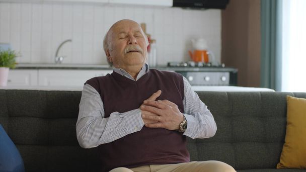 Sensation d'inconfort, risque de crise cardiaque. Homme âgé ressentant une douleur thoracique alors qu'il était assis sur le canapé à la maison ayant des difficultés à respirer. Il touche sa poitrine avec sa main. - Photo, image