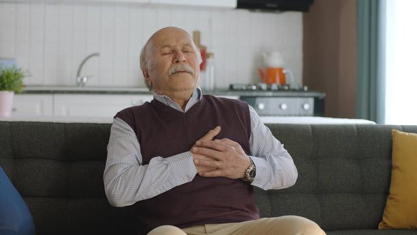 Epämukavuuden tunne, sydänkohtauksen riski. Iäkäs mies tunne rintakipua istuessaan sohvalla kotona, joilla on hengitysvaikeuksia. Hän koskettaa rintaansa kädellään.. - Valokuva, kuva