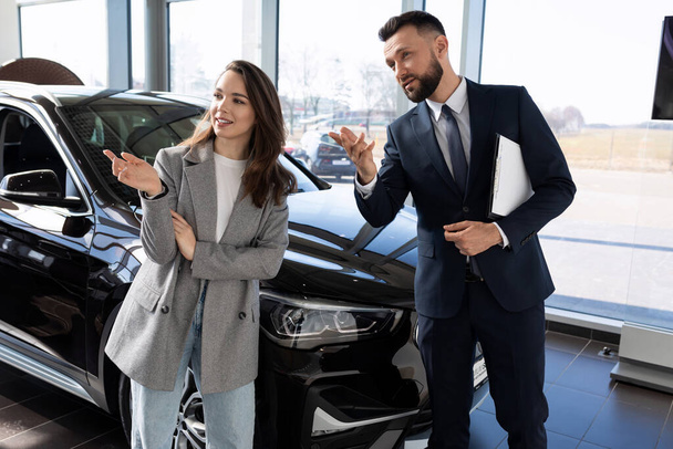 eine junge Frau in einem Autohaus, zusammen mit einem Mitarbeiter, ein Autohändler wählt ein neues Auto. - Foto, Bild