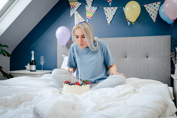 Happy woman in pajama and party cap blowing out candles on birthday cake on the bed in decorated bedroom. Make a wish process. Morning surprise. Happy birthday concept. Selective focus - Foto, afbeelding