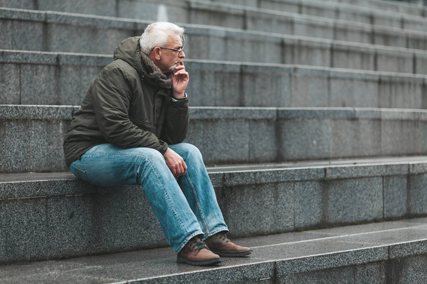 Gray-haired man is lost in thought and sits on the steps. Nostalgia for a pensioner. - Foto, Imagen