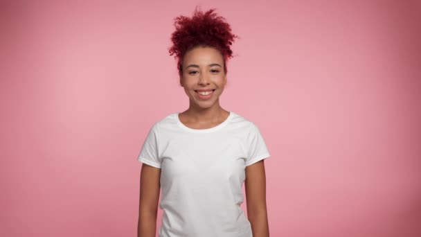 Hymyilevä afroamerikkalainen punapää kihara nainen valkoisessa t-paidassa poseeraa kääntyy pää ja näyttää kameralta. Muotokuva positiivinen Afrikkalainen nainen seisoo eristetyllä vaaleanpunaisella taustalla. Ihmiset tunteet elämäntapa - Materiaali, video
