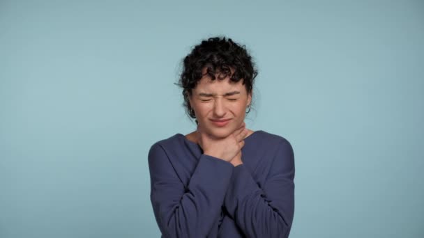 Unhealthy young curly woman in blue sweater touching neck feeling discomfort or pain. Sick sad beautiful female suffering from angina, sore throat or loss of voice on isolated light blue background - Filmagem, Vídeo