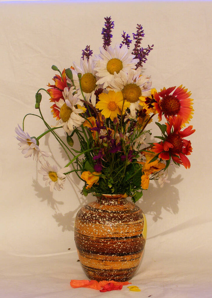 Μπουκέτο με καλοκαιρινά λουλούδια σε πήλινο βάζο σε ελαφρύ υφασμάτινο φόντο. Ροδάκινα στέκεται σε ένα βαθύ πιάτο δίπλα στο άλλο - Φωτογραφία, εικόνα