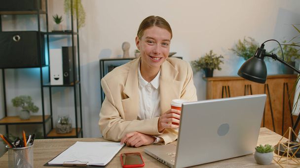 Businesswoman freelancer enters office start working on laptop computer at desk and drinking morning coffee. Woman works on notebook, sends messages makes online purchases watching social media movies - Fotoğraf, Görsel