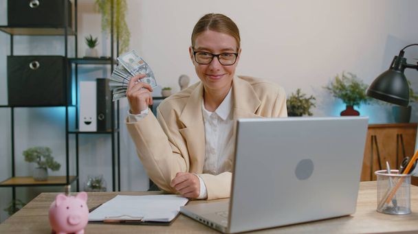 Happy businesswoman working on laptop computer sincerely rejoicing win, receiving money dollar cash banknotes, success lottery luck at office workplace. Professional manager freelancer woman dancing - 写真・画像